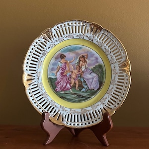 May include: A white porcelain plate with a yellow rim and gold trim. The plate features a delicate openwork design and a central image of three figures in a landscape setting.