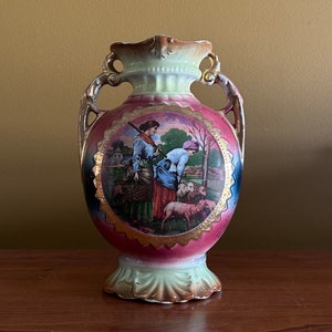 May include: A vintage porcelain vase with two women and sheep painted on it. The vase has gold accents and a pink and green glaze.