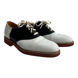 May include: A pair of white and black leather saddle shoes with white laces. The shoes have a red rubber sole.