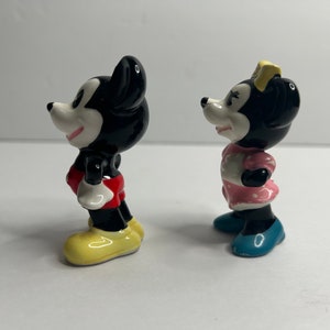 Może przedstawiać: Para ceramicznych figurek przedstawiających Myszki Miki i Minnie. Miki ma na sobie czerwone spodenki i ż&oacute;łte buty. Minnie ma na sobie r&oacute;żową sukienkę w białe kropki i niebieskie buty.