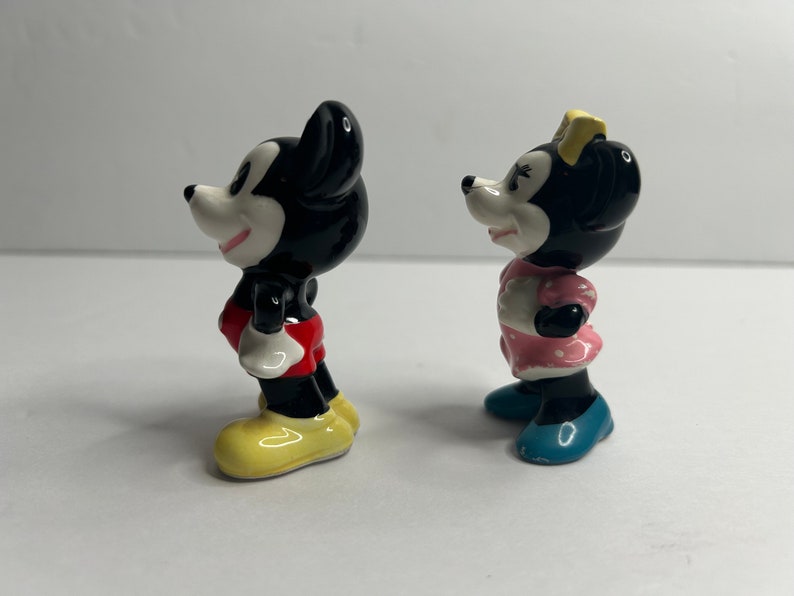Może przedstawiać: Para ceramicznych figurek przedstawiających Myszki Miki i Minnie. Miki ma na sobie czerwone spodenki i ż&oacute;łte buty. Minnie ma na sobie r&oacute;żową sukienkę w białe kropki i niebieskie buty.