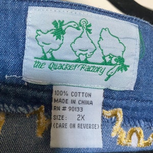 May include: A blue denim fabric with a white tag that says "The Quacker Factory" with a green border and a cartoon image of two ducks. The tag also says "100% Cotton, Made in China, RN # 90133, Size: 2X, (Care on Reverse)".