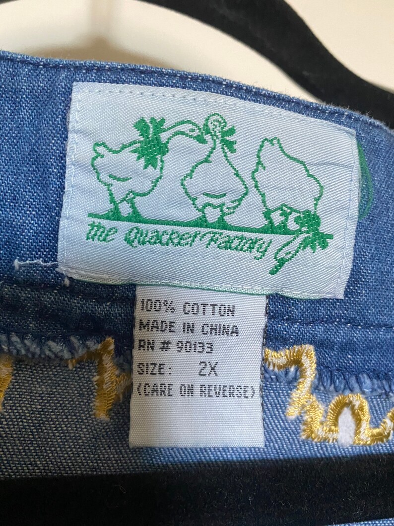 May include: A blue denim fabric with a white tag that says "The Quacker Factory" with a green border and a cartoon image of two ducks. The tag also says "100% Cotton, Made in China, RN # 90133, Size: 2X, (Care on Reverse)".
