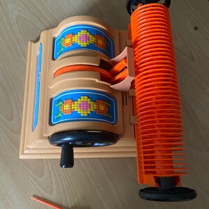 May include: A vintage plastic toy loom with orange and black spools and a wooden base. The loom has a crank handle and a pattern of colorful squares on the front.