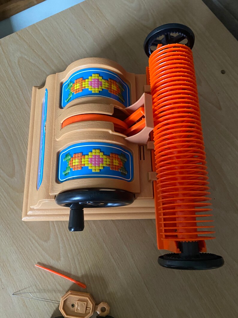 May include: A vintage plastic toy loom with orange and black spools and a wooden base. The loom has a crank handle and a pattern of colorful squares on the front.