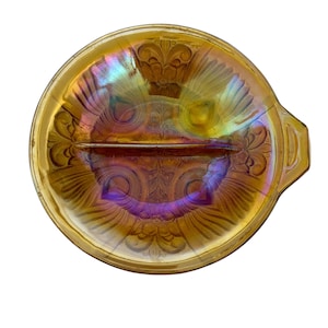 May include: An iridescent, amber-colored divided serving dish with a decorative floral pattern. The dish has a handle on one side and a raised divider in the center. The dish is round and has a scalloped edge.