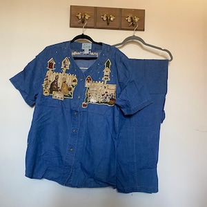 May include: A blue denim shirt and pants set with a colorful embroidered design of a house, flowers, and animals. The shirt has a button-up front and short sleeves.