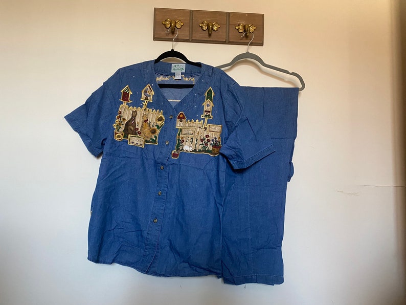 May include: A blue denim shirt and pants set with a colorful embroidered design of a house, flowers, and animals. The shirt has a button-up front and short sleeves.