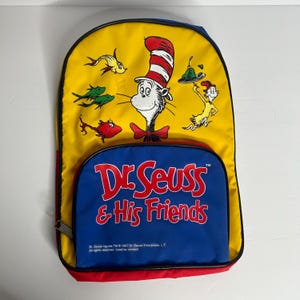 May include: Yellow backpack featuring Dr. Seuss characters, including the Cat in the Hat and fish. The backpack has a blue pocket with the text "Dr. Seuss & His Friends" in red. The backpack is trimmed in red and black.