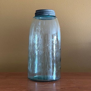 Op de afbeelding: Een vintage blauw getinte glazen pot met een metalen deksel. De pot heeft een brede opening en een afgeronde body, met het woord "Ball" in reliëf op de voorkant. De pot wordt waarschijnlijk gebruikt voor het inmaken of bewaren.