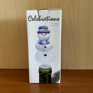May include: A 6-inch snowman wine cork in a box. The snowman has a blue hat and scarf, with black button eyes and a carrot nose. The box reads "Celebrations" and "6" Snowman Wine Cork."