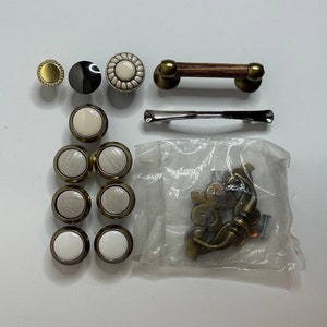 May include: Assortment of cabinet hardware including knobs and pulls in various styles and finishes. There are round knobs with a white ceramic finish, round knobs with a wood grain finish, a round knob with a black finish, a round knob with a gold finish, a gold pull with a wood handle, a silver pull, and a decorative gold pull with a scroll design.