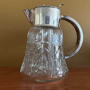 May include: A clear glass pitcher with a silver lid and handle. The pitcher has a cut glass design with floral patterns.