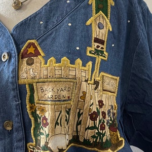 May include: A blue denim jacket with a colorful embroidered patch depicting a backyard garden scene. The patch features a white picket fence, flowers, a birdhouse, a rabbit, and a sign that reads "Back Yard Garden".