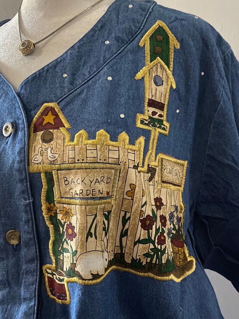 May include: A blue denim jacket with a colorful embroidered patch depicting a backyard garden scene. The patch features a white picket fence, flowers, a birdhouse, a rabbit, and a sign that reads "Back Yard Garden".