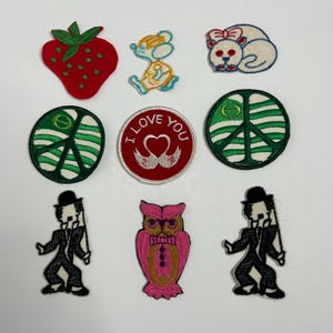 May include: A collection of embroidered patches featuring various designs. Includes a red strawberry, a mouse, a white cat with a bow, peace signs, a red patch with "I Love You" text, an owl, and figures in suits.