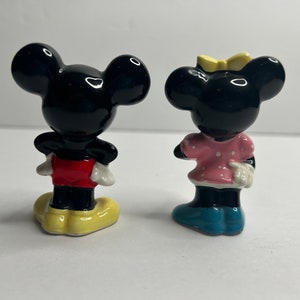 Może przedstawiać: Para ceramicznych figurek przedstawiających Myszki Miki i Minnie. Miki ma na sobie czerwone spodenki i ż&oacute;łte buty. Minnie ma na sobie r&oacute;żową sukienkę w białe kropki i niebieskie buty.