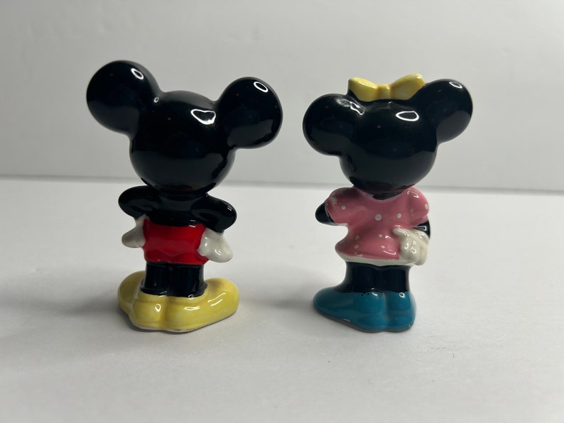 Może przedstawiać: Para ceramicznych figurek przedstawiających Myszki Miki i Minnie. Miki ma na sobie czerwone spodenki i ż&oacute;łte buty. Minnie ma na sobie r&oacute;żową sukienkę w białe kropki i niebieskie buty.