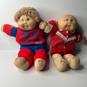 May include: Two vintage Cabbage Patch Kids dolls. One doll wears a red and blue outfit with the number 31 on the shirt. The other doll is bald and wears a red plaid outfit. Both dolls have soft, fabric bodies and plastic faces.