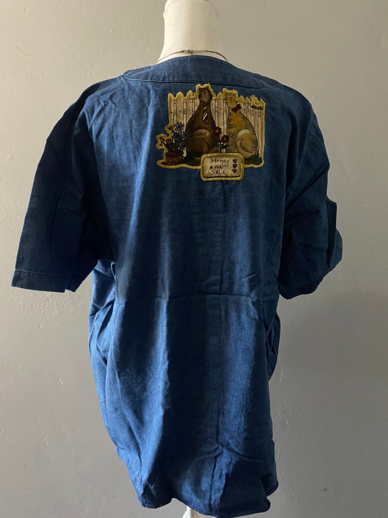 May include: A blue denim shirt with a patch on the back featuring two cats sitting by a fence with the text "Honey, Good Sale".