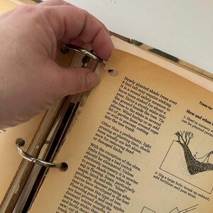 May include: Open vintage book with text about planting and pruning trees. The book is open to a page with text and illustrations. A hand is holding the metal rings of the binder.