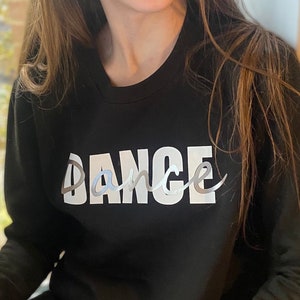 May include: Black crew neck sweatshirt with the word "DANCE" printed across the front in a white, silver, and light blue design. The sweatshirt is made of a soft material.