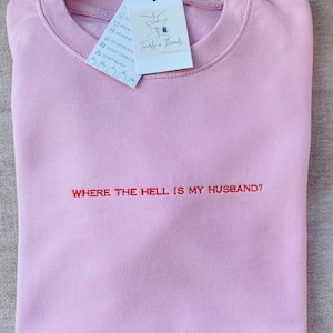 May include: Pale pink sweatshirt with the embroidered phrase "WHERE THE HELL IS MY HUSBAND?" in red. A tag from "Twirls & Threads" is attached. The garment is neatly folded.