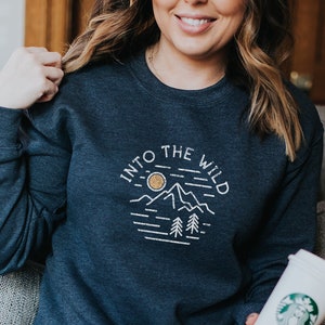 May include: A dark heather grey sweatshirt with a white graphic design that says "Into the Wild" with a mountain and sun illustration.