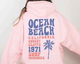 May include: Light pink adult unisex hoodie with a retro graphic design on the back. The design features the text "Ocean Beach California Sunset Cliffs 1971 Surf Memories" in a blue and red font with a palm tree graphic.