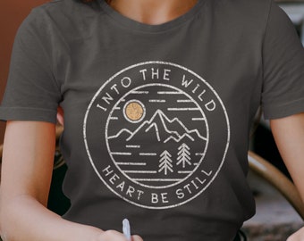 May include: A woman wearing a gray t-shirt with a white graphic design that says "Into the Wild Heart Be Still" and a mountain scene with a sun and trees.
