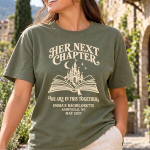 May include: An olive green t-shirt featuring a cream-colored graphic of an open book with a castle rising from the pages, a crescent moon, and stars. The text reads: HER NEXT CHAPTER, WE ARE IN THIS TOGETHER, EMMA'S BACHELORETTE, ASHEVILLE, NC, MAY 2027.