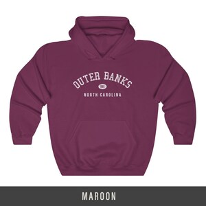 Puede incluir: Sudadera con capucha color granate con el texto "Outer Banks" en blanco, con un peque&ntilde;o c&iacute;rculo que contiene el texto "OBX" debajo. El texto "North Carolina" est&aacute; debajo del c&iacute;rculo.