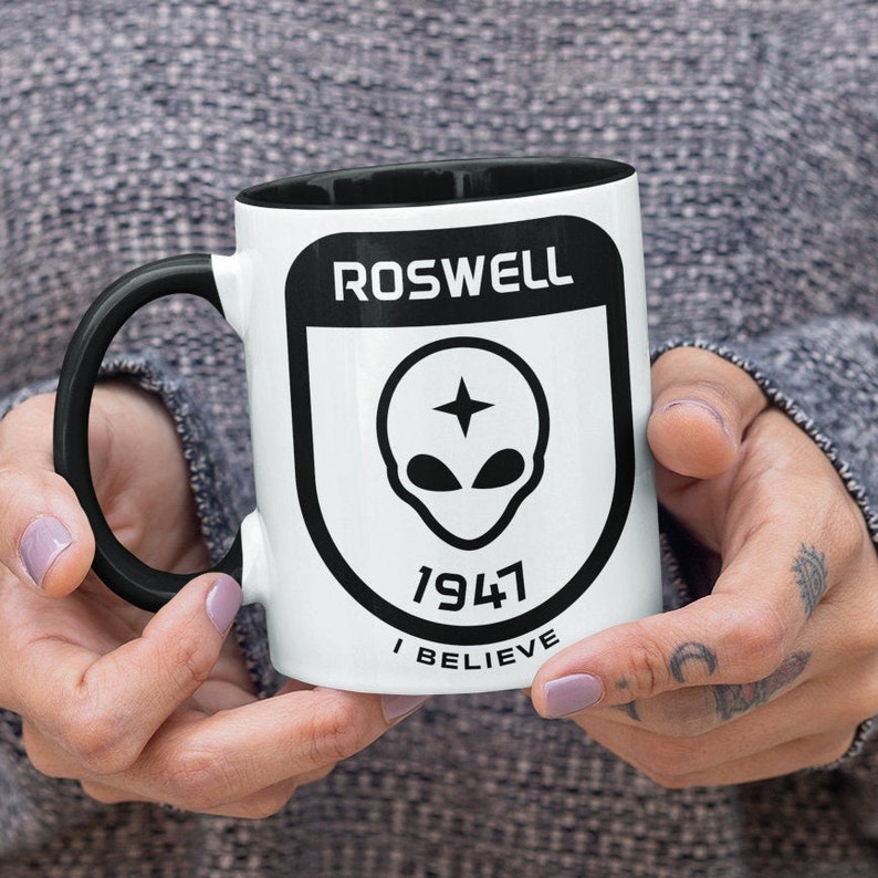 Pu&ograve; includere: Una tazza in ceramica bianca con manico e bordo neri. La tazza presenta una grafica in bianco e nero di una testa aliena con una stella sulla fronte all'interno di uno scudo con il testo "Roswell 1947 I Believe".