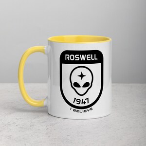Pu&ograve; includere: Una tazza in ceramica bianca con un bordo giallo. La tazza presenta una grafica nera di una testa aliena con una stella sulla fronte all'interno di uno scudo con il testo "Roswell 1947 I Believe".