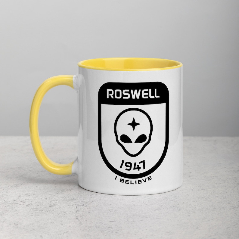 Pu&ograve; includere: Una tazza in ceramica bianca con un bordo giallo. La tazza presenta una grafica nera di una testa aliena con una stella sulla fronte all'interno di uno scudo con il testo "Roswell 1947 I Believe".