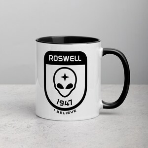 Pu&ograve; includere: Una tazza in ceramica bianca con bordo e manico neri. La tazza presenta una grafica in bianco e nero di una testa aliena con una stella sopra, circondata da una forma a scudo. Il testo "ROSWELL 1947 I BELIEVE" &egrave; stampato sotto la testa aliena.