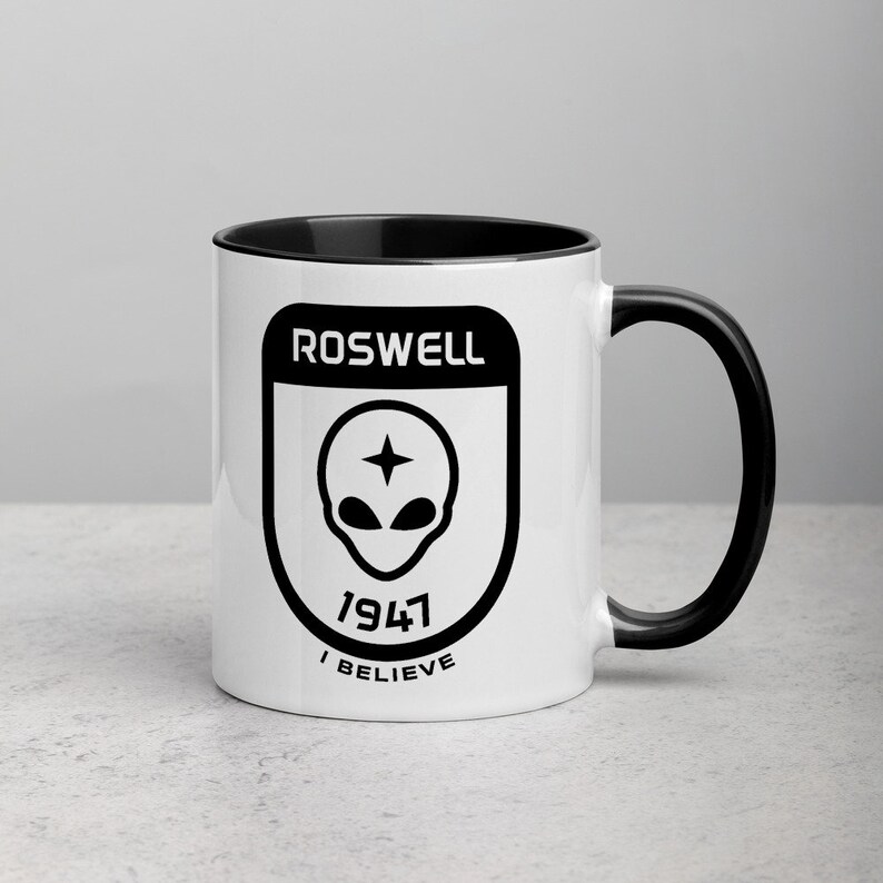 Pu&ograve; includere: Una tazza in ceramica bianca con bordo e manico neri. La tazza presenta una grafica in bianco e nero di una testa aliena con una stella sopra, circondata da una forma a scudo. Il testo "ROSWELL 1947 I BELIEVE" &egrave; stampato sotto la testa aliena.