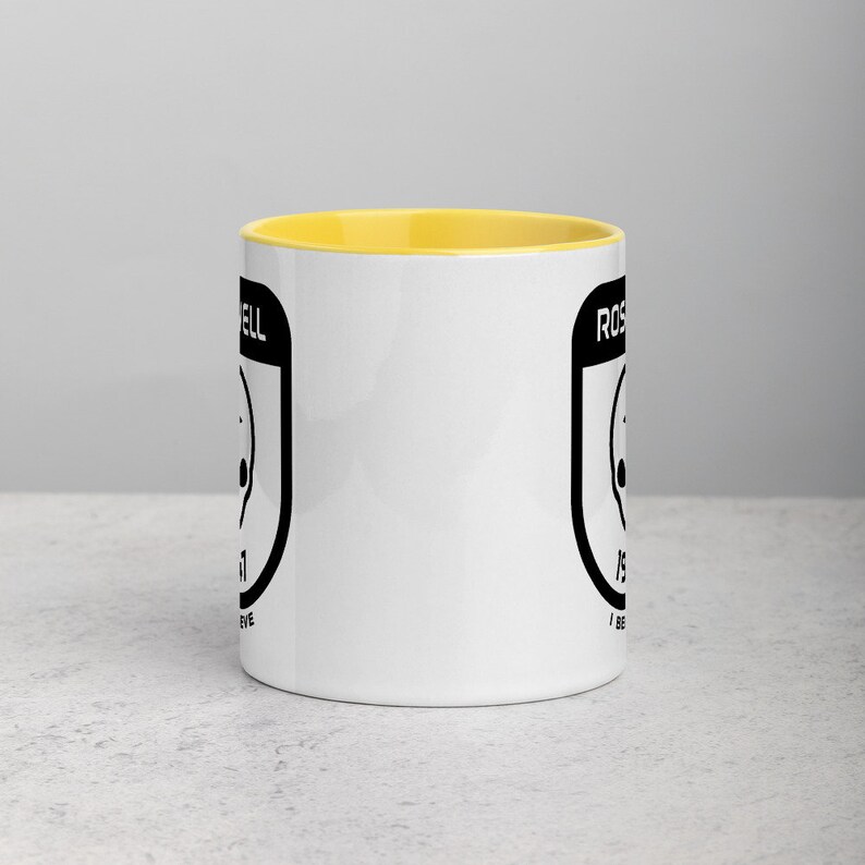 Pu&ograve; includere: Tazza in ceramica bianca con bordo giallo. La tazza presenta una grafica nera di un UFO con il testo "Roswell" e "Io credo" su entrambi i lati.