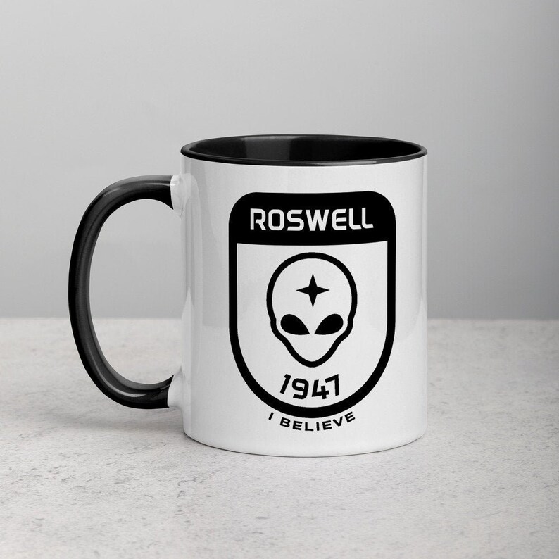 Pu&ograve; includere: Una tazza in ceramica bianca con manico e bordo neri. La tazza presenta una grafica in bianco e nero di una testa aliena con una stella sulla fronte all'interno di uno scudo con il testo "Roswell 1947 I Believe".