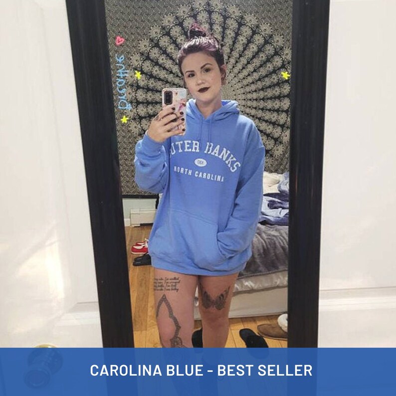 Puede incluir: Una sudadera con capucha azul claro con el texto "Outer Banks" y "North Carolina" en blanco. La sudadera la lleva puesta una persona en un reflejo de espejo.