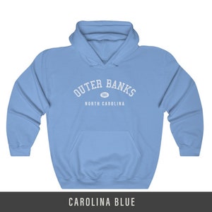 Puede incluir: Una sudadera con capucha azul cielo con el texto "Outer Banks" en blanco, con un peque&ntilde;o c&iacute;rculo que contiene el texto "OBX" debajo y el texto "North Carolina" debajo.