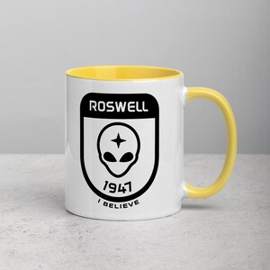 Pu&ograve; includere: Tazza in ceramica bianca con bordo e manico gialli. La tazza presenta una grafica in bianco e nero di una testa aliena con una stella sopra, circondata da uno scudo nero con il testo "ROSWELL 1947 I BELIEVE".