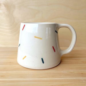 Puede incluir: Una taza de cerámica blanca con un acabado moteado y un asa curvada. La taza está decorada con guiones verticales de colores en rojo, amarillo y verde. La taza está sobre un fondo de madera clara.