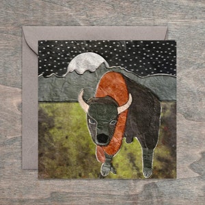 Puede incluir: Un bisonte negro y naranja con cuernos blancos se encuentra en un campo con una luna llena y estrellas en el fondo. La imagen está hecha de papel y tiene un aspecto texturizado.