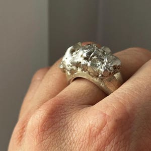 Könnte beinhalten: Ein silberner Ring mit einem strukturierten, organischen Design. Die Oberfläche des Rings hat ein raues, kristallines Aussehen mit mehreren Facetten und unregelmäßigen Formen. Das Band ist glatt und breit und steht im Kontrast zur strukturierten Oberseite.