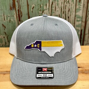 May include: Gray and white trucker hat with a North Carolina state outline patch in purple, yellow, and white. The patch has the letters "N*C" in purple. The hat has a black label with the text "Richardson" and "112" on it.