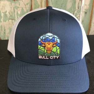 May include: A navy blue and white trucker hat with a stained glass bull design and the text "BULL CITY" on the front.