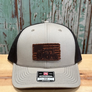 May include: A gray and black trucker hat with a brown leather patch featuring an American flag with a tractor on it. The hat has the text "Richardson" and "112" on the front.