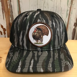 May include: A black and green camouflage trucker hat with a brown circular patch featuring a detailed embroidered image of a wild turkey.