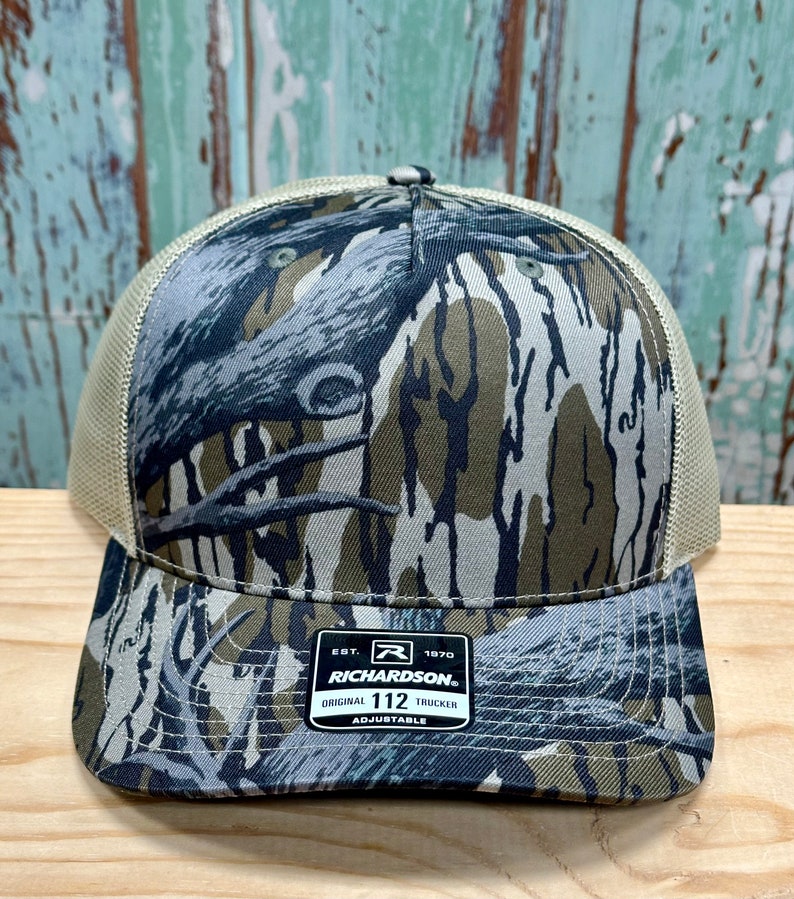 May include: A camouflage trucker hat with a mesh back and a curved peak. The hat features a Richardson 112 label and is adjustable. The camouflage design incorporates shades of green, brown, and grey.